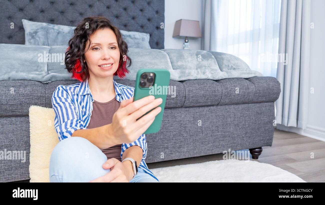 Eine Frau mit lockigem Haar genießt ihr Smartphone, während sie in einem stilvollen Wohnzimmer auf dem Boden sitzt und einen modernen Lebensstil und Komfort widerspiegelt Stockfoto