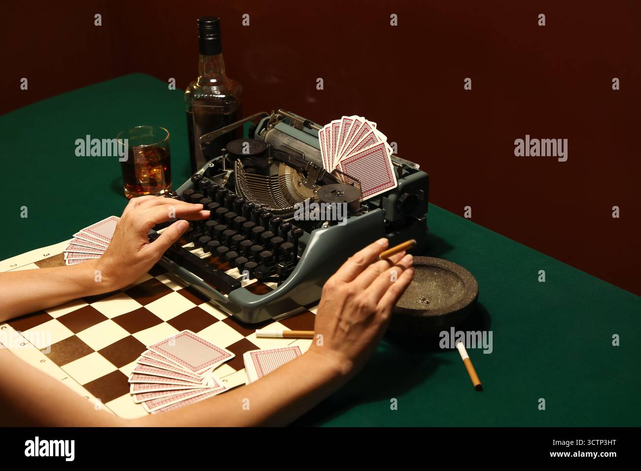 Junge Frau mit Vintage-Schreibmaschine, Spielkarten und Zigarette am Tisch auf grünem Tisch in der Nähe der braunen Wand Stockfoto