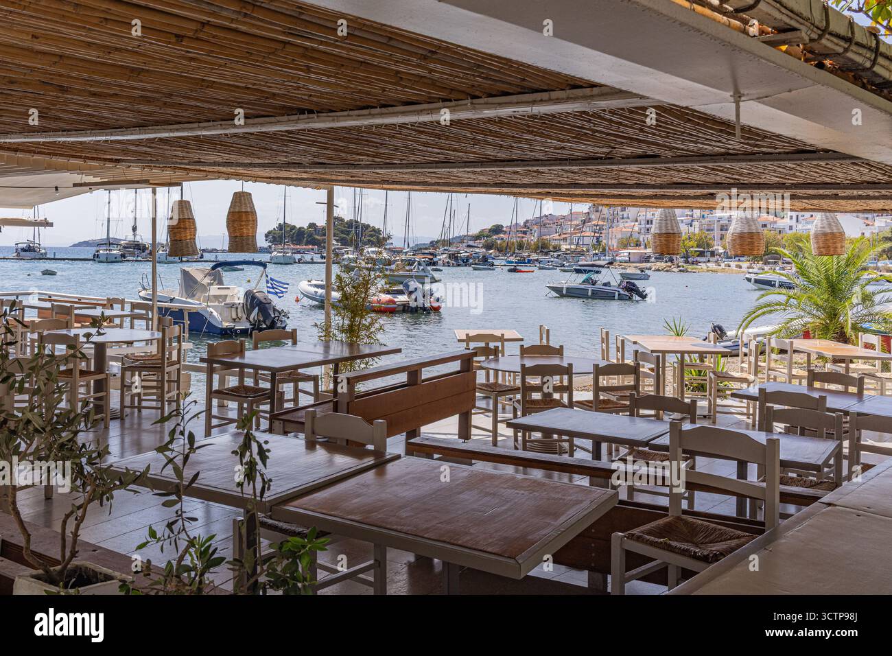 Taverne am Meer mit Blick auf den Hafen von Skiathos Island, Griechenland. Holztische unter einer schattigen Terrasse blicken auf das ruhige türkisfarbene Wasser mit verankerten Booten. Stockfoto