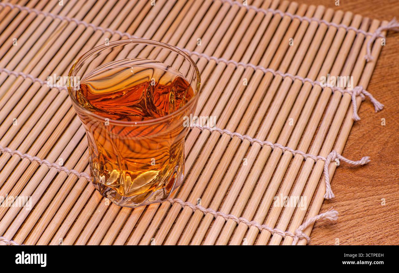 Ein klares Glas gefüllt mit Getränk auf einer Bambusmatte über einer Holzoberfläche. Die warme bernsteinfarbene Farbe des Getränks ist ein wunderschöner Kontrast. Stockfoto