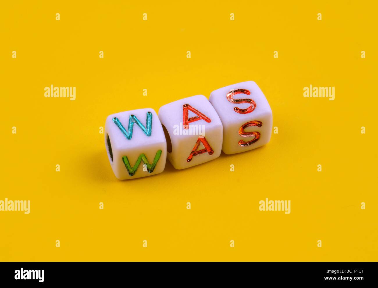 Drei weiße Würfel mit farbenfrohen Gravurbuchstaben, die das Wort „WAS“ schreiben, auf einem hellgelben Hintergrund platziert. Die verspielten Würfel symbolisieren Sprache. Stockfoto