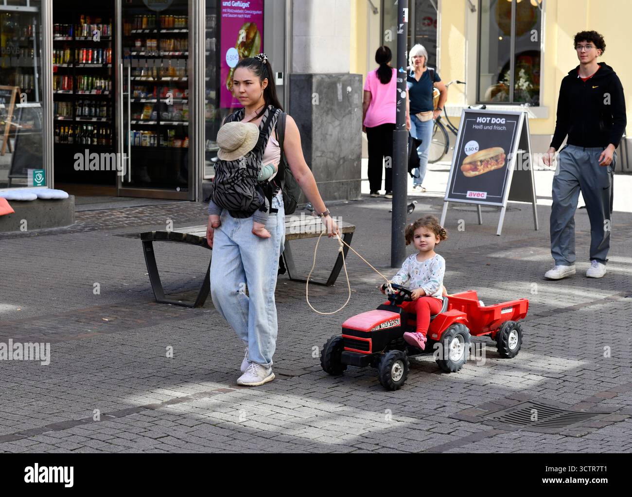 Frau, die ein Baby trägt und ein Kind auf einem Spielzeugtraktor zieht Heidelberg in Südwestdeutschland. Stockfoto