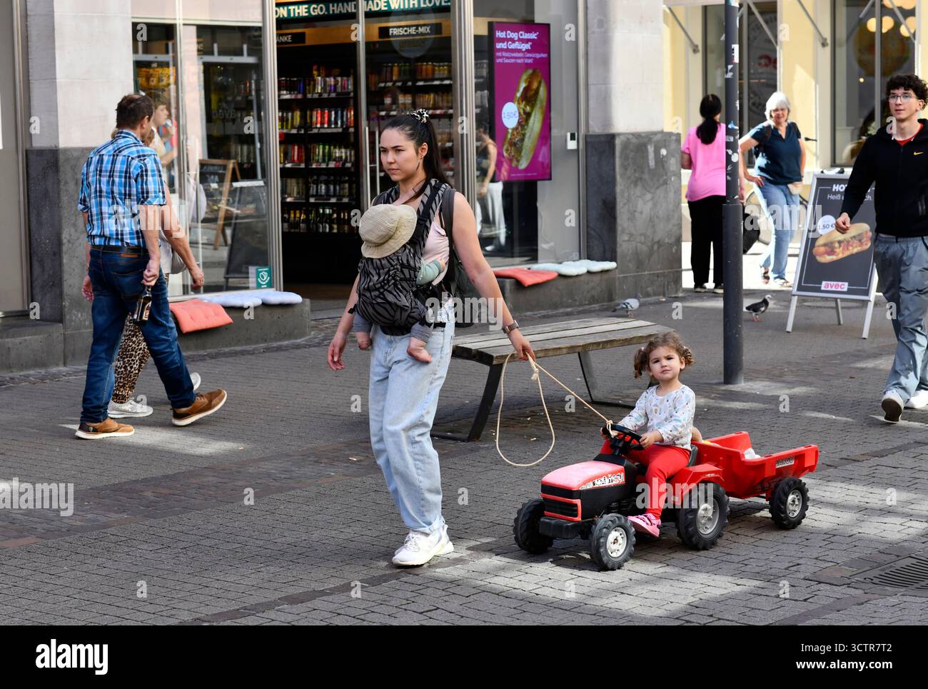 Frau, die ein Baby trägt und ein Kind auf einem Spielzeugtraktor zieht Heidelberg in Südwestdeutschland. Stockfoto