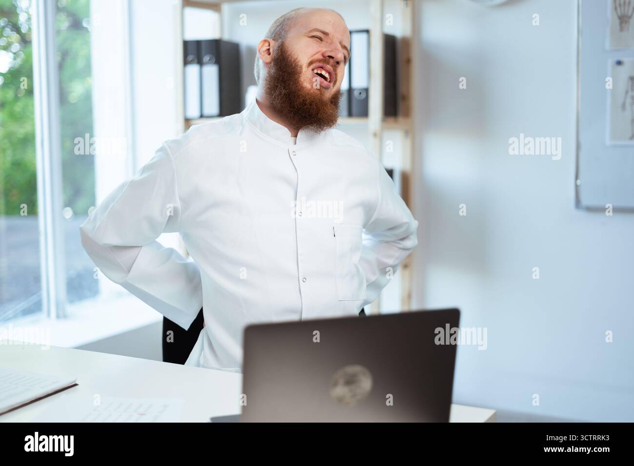 Ein bärtiger Mann Arzt, der an Rückenschmerzen, Lendenwirbelproblemen und Wirbelsäulenverletzungen durch sitzende Arbeit leidet. Stockfoto
