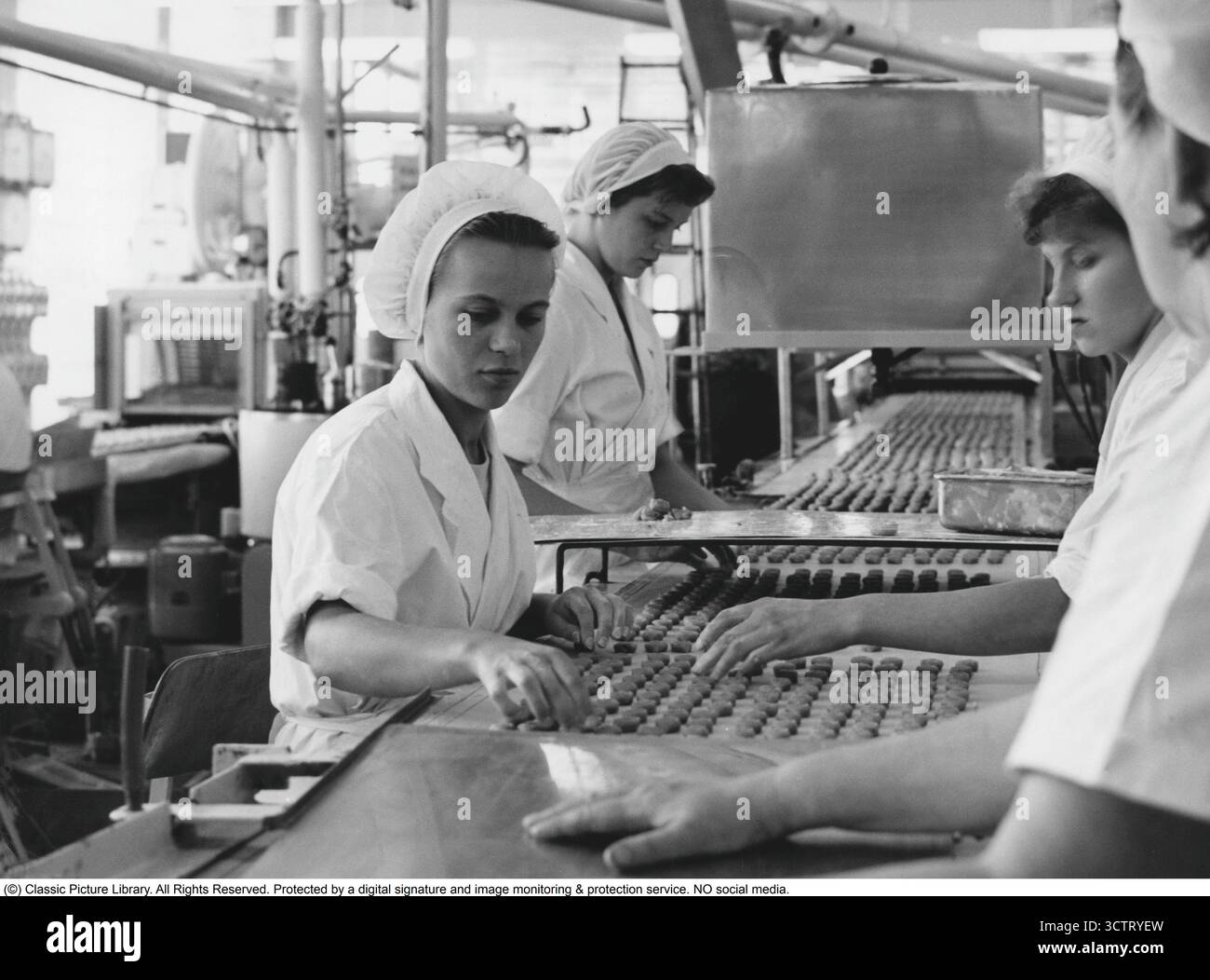 Ab Marabou, Sundbyberg 1957. Frauen sitzen an einem Förderband, während Marabous Marzipanpralinen ihre glänzende Schokoladenbeschichtung in der so genannten Enrobing-Maschine erhalten. Rund eine halbe Million Pralinen passieren in jeder Schicht entlang der Linie. Marabou wurde 1916 gegründet und entwickelte sich schnell zu einem der beliebtesten Schokoladenproduzenten Schwedens. Das Werk in Sundbyberg, kurz vor den Toren Stockholms, war jahrzehntelang der Hauptproduktionsstandort. In den 1950er Jahren setzte sich Marabou für die Herstellung von Süßwaren im industriellen Maßstab ein, wobei Präzisionsmaschinen mit handwerklicher Arbeit kombiniert wurden, die oft von Frauen ausgeführt wurde. Stockfoto