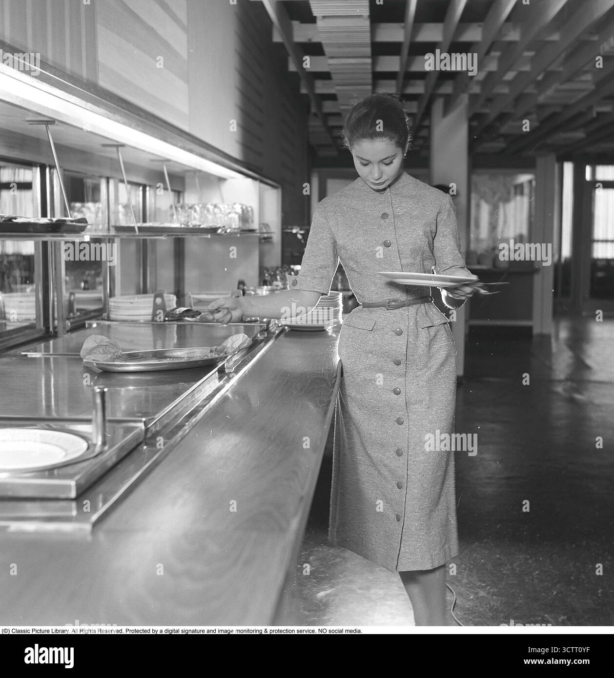 Sie kleidete sich schön 1956. Das junge Model hilft sich beim Essen im Restaurant. Sie trägt die typische Kleidung der Periode, die Hemdkleid oder Hemdkleid genannt wird. November 1956 Stockfoto