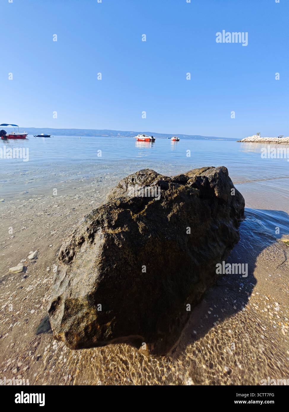 Ein großer Felsen liegt am Sandstrand, während kleine Boote an einem sonnigen Tag mit klarem, blauem Himmel im klaren, ruhigen Wasser in der Nähe der Küste schweben. Stockfoto