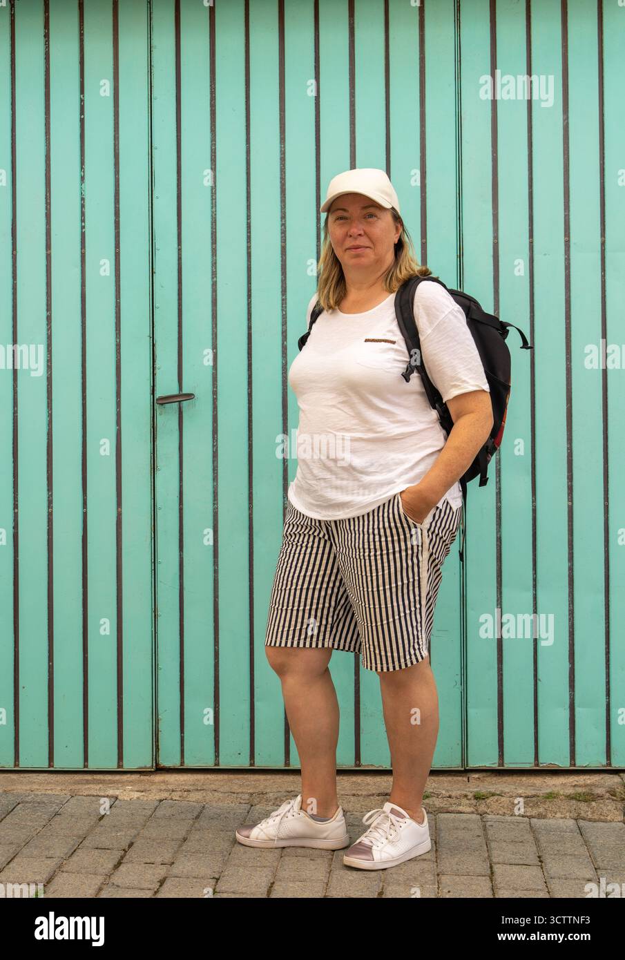 Porträt einer Frau mittleren Alters in Baseballmütze, die vor dem verwitterten blauen Holztor steht. Lässiger Outdoor-Look, Tageslicht. Echte Menschen, authen Stockfoto