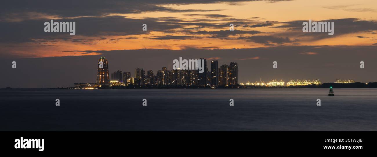 Weit entfernte Skyline von Forest City, Johor, Malaysia, von der Westküste Singapurs in der Abenddämmerung aus gesehen. Beleuchtete Gebäude und Hafenkräne sichtbar. Stockfoto