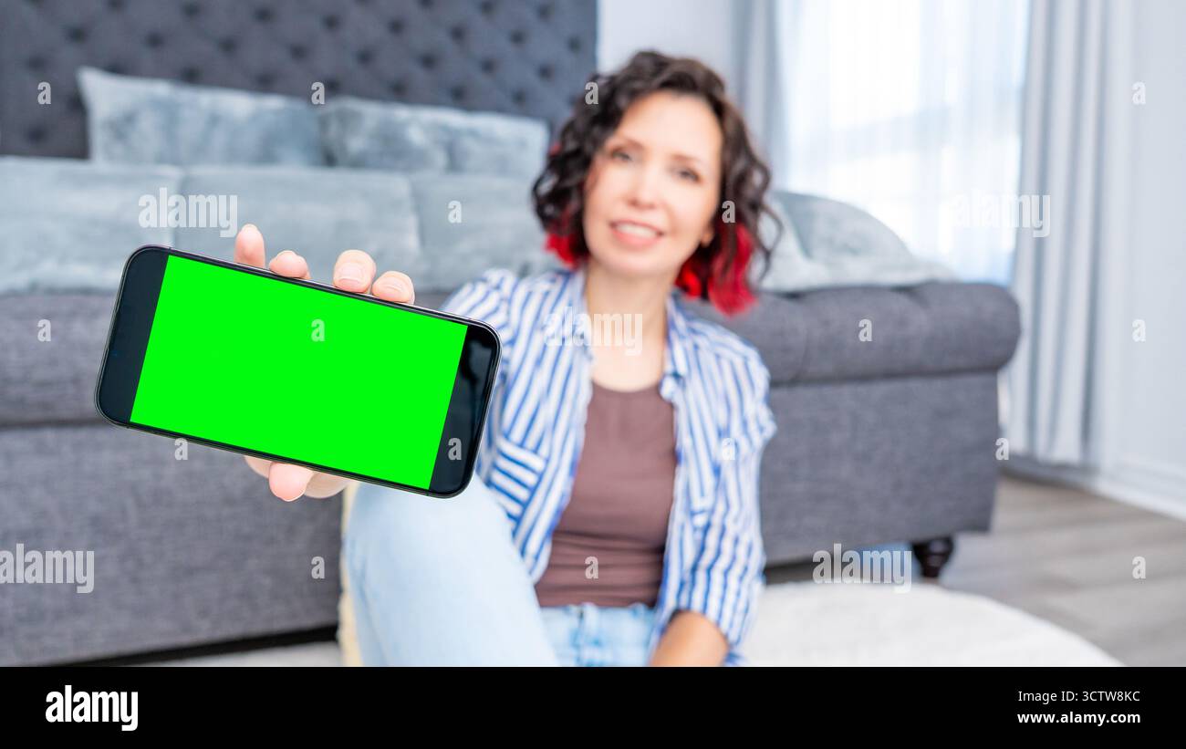 Frau zeigt Smartphone-Mockup mit grünem Bildschirm, während sie auf dem Boden in einem stilvollen Wohnzimmer sitzt und eine komfortable und einladende Atmosphäre hervorhebt Stockfoto