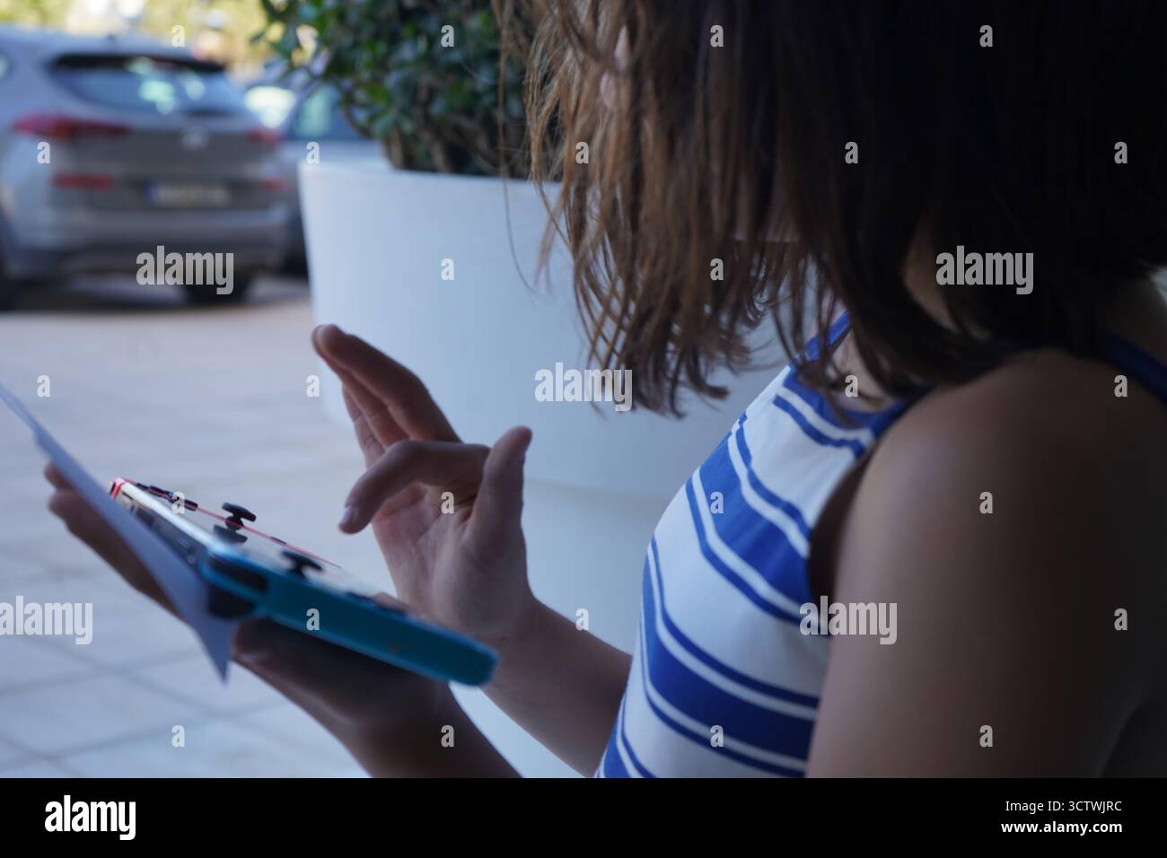 Eine Nahaufnahme eines Mädchens, das im Freien eine Nintendo Switch-Handspielkonsole spielt, während er eine kleine Note mit blau-weißem Oberteil hält. Stockfoto