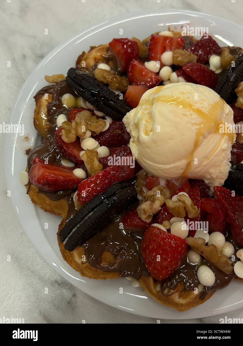 Das Bild zeigt eine Nahaufnahme eines Desserttellers mit einer Waffel bedeckt mit Schokoladenaufstrich, frischen Erdbeeren, Oreo-Keksen, Walnüssen und weißer Schokolade Stockfoto