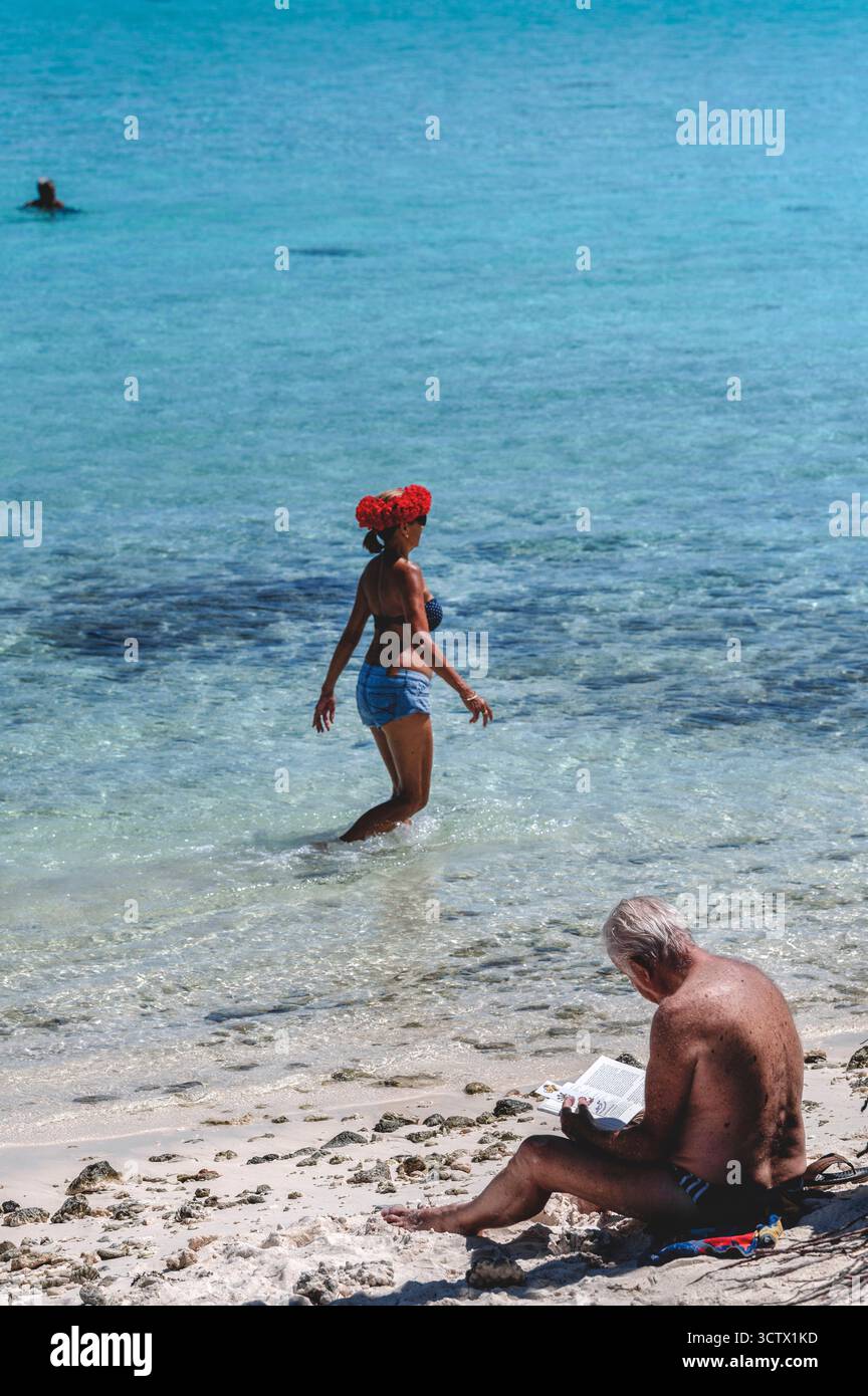 Frau, die in flachem türkisfarbenem Wasser in einer roten Kopfhülle läuft und während ein Mann am Strand vergessen liest, ein ehrlicher Moment unter tropischem Sonnenlicht. Stockfoto