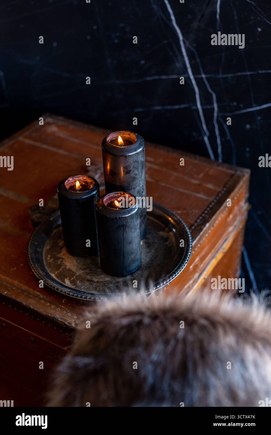 Schwarze Kerzen brennen Flamme auf silberner Zinnplatte auf altem Lederstamm mit Pelz im Vordergrund Stockfoto