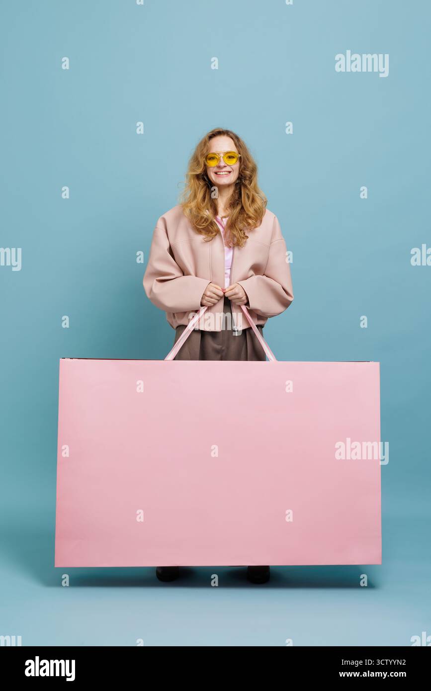 Glücklicher Käufer mit einer übergroßen rosa Tasche vor blauem Studiohintergrund, ideal für Black Friday, Rabatte, Werbeanzeigen im Einzelhandel, Banner und soziale Kampagnen. Stockfoto