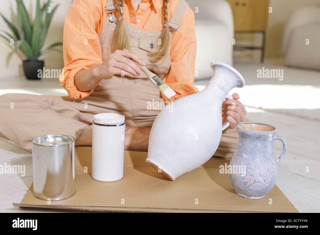 Nahaufnahme einer Frau, die eine strukturierte Keramikvase mit weißer Kreidefarbe neu malte, zeigt Heimwerker und umweltfreundliche Wohnkultur bei hellem Tageslicht. Stockfoto
