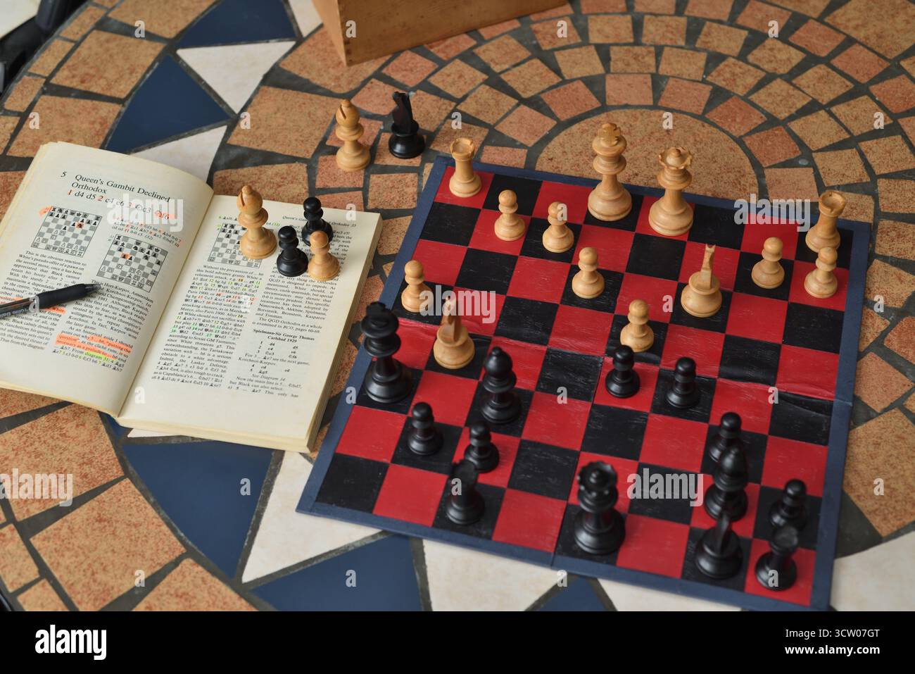 Schachbuch und Vintage-Schachset mit Boxholz Staunton-Schachfiguren auf einem roten und schwarzen Brett, die das Queens Gambit-Schach erlernen, das den alten Weg eröffnet. Stockfoto