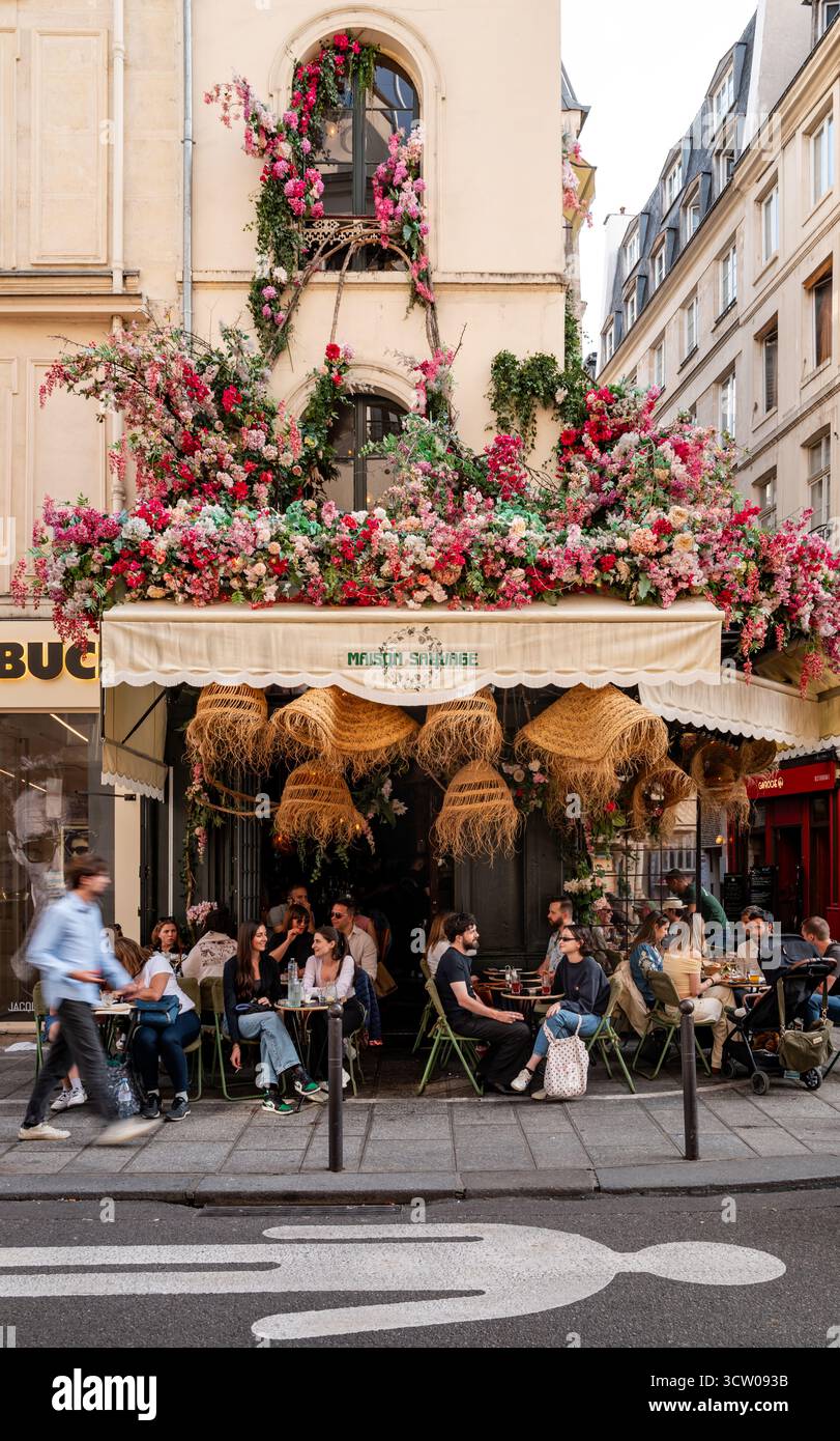 Das Pariser Straßencafé ist mit blühenden Blumen bedeckt und an einem sonnigen Tag treffen sich die Menschen und speisen im Freien. Stockfoto