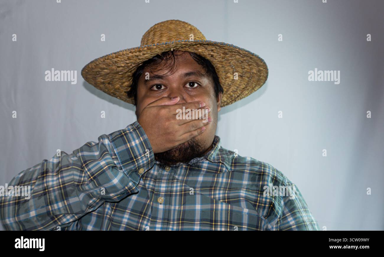 Ein indonesischer Mann trägt einen Strohhut und ein grünes kariertes Hemd auf weißem Hintergrund. Die Hand bedeckt den Mund Stockfoto