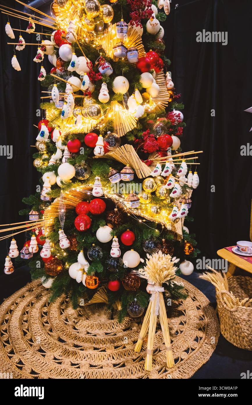 Majestätisch steht ein atemberaubender Weihnachtsbaum, der mit einer Vielzahl einzigartiger Ornamente und leuchtenden Lichterketten geschmückt ist. Die Dekorationen umfassen cl Stockfoto