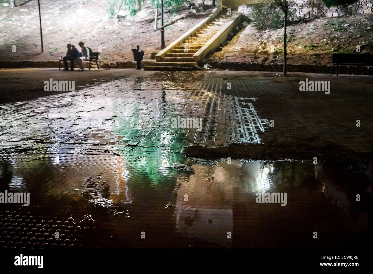 Aus nächster Nähe ein Blick auf einen nassen Kopfsteinpflasterplatz in Villajoyosa mit einer großen Pfütze, die die hellen Lichter und Farben des Ar reflektiert Stockfoto