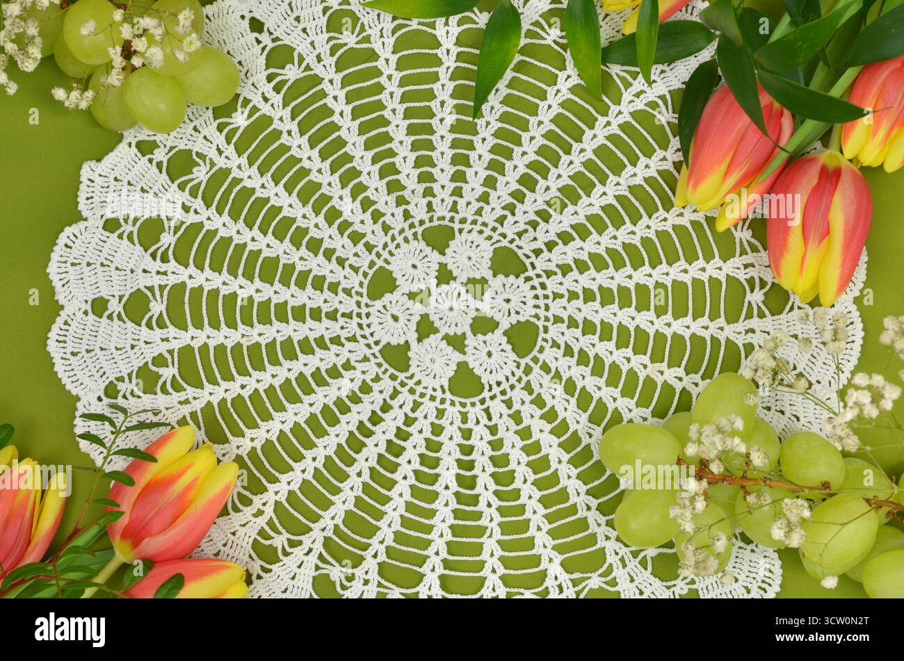 Draufsicht: Retro, handgefertigtes Deckchen auf grünem Hintergrund mit Blumen: Rote, gelbe Tulpen und weiße gypsophila, grüne Trauben. Stockfoto