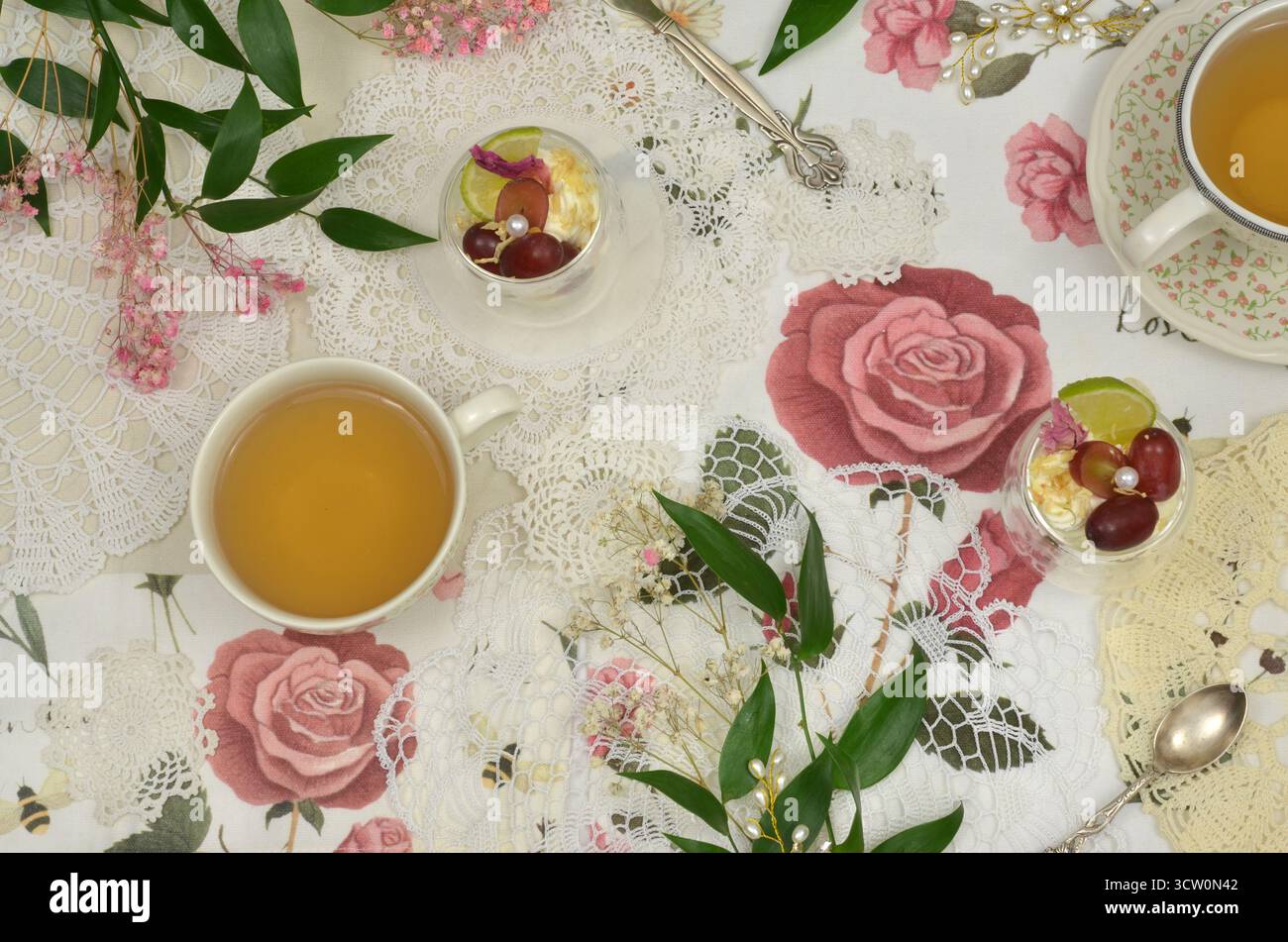 Retro-Tisch mit Blick von oben, dekoriert mit einer Tischdecke mit Rosen, Häkeldecken und grünen Blättern. Zwei Tassen Tee und Obstdesserts. Stockfoto