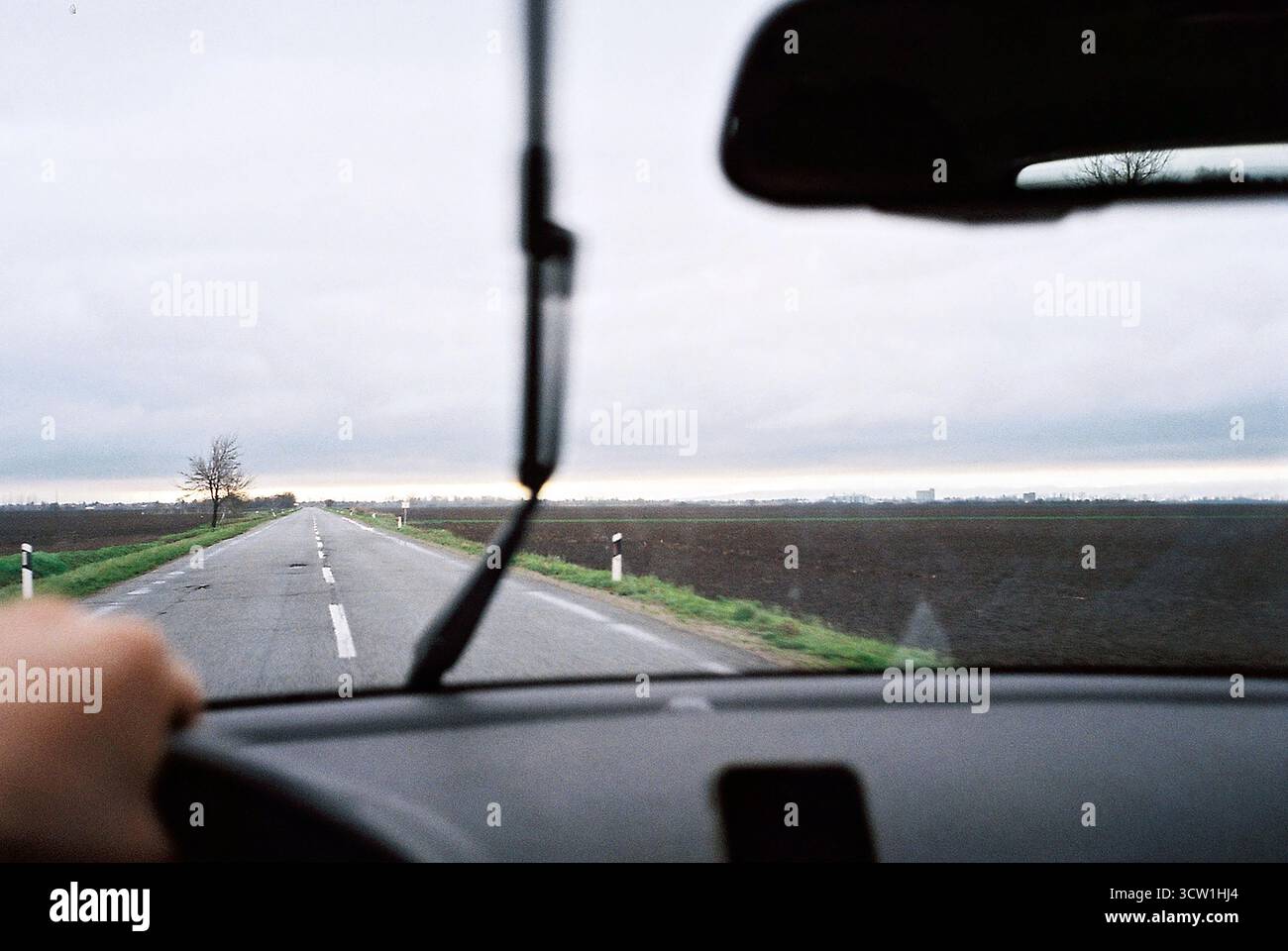Analoges 35-mm-Foto aus Sicht des Fahrers bei leichtem Regen. Ein Scheibenwischer durchschneidet Regentropfen und offenbart ein gepflügtes Feld unter bewölktem Himmel. Stockfoto