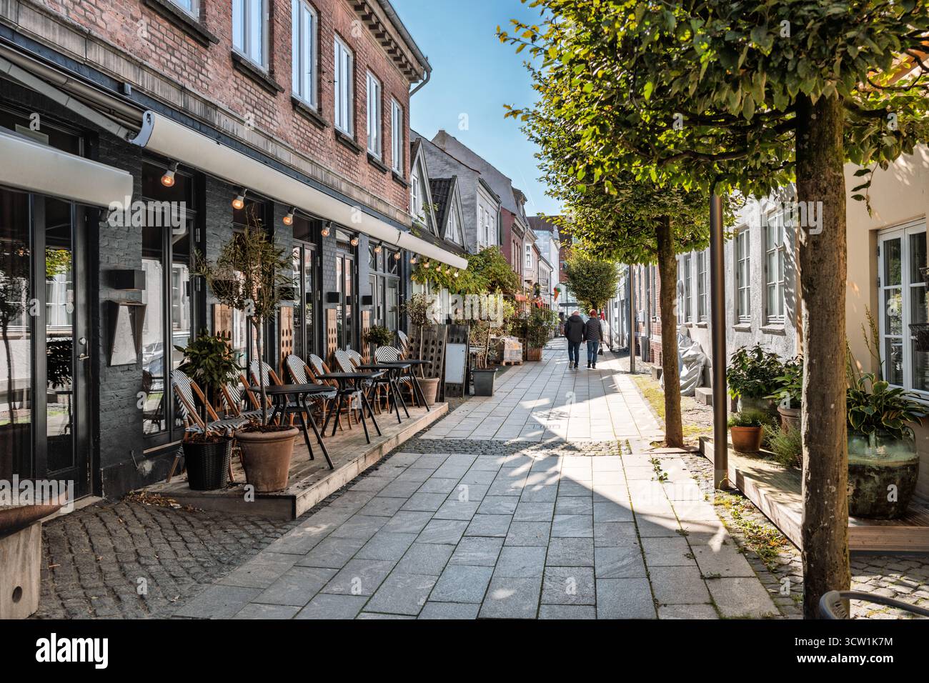 Eine sonnige Straßenszene mit einer Reihe von Cafés vor Backsteinfronten, Topfpflanzen und Fußgängern, die auf einer gepflasterten Fußgängerzone spazieren. A wa Stockfoto