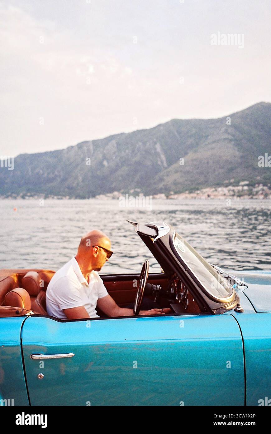 Analoges 35-mm-Foto eines Glatzen-Mannes mit Sonnenbrille in einem Vintage-Cabrio auf einem Steindock bei Sonnenuntergang, mit Bucht von Kotor im Hintergrund. Stockfoto