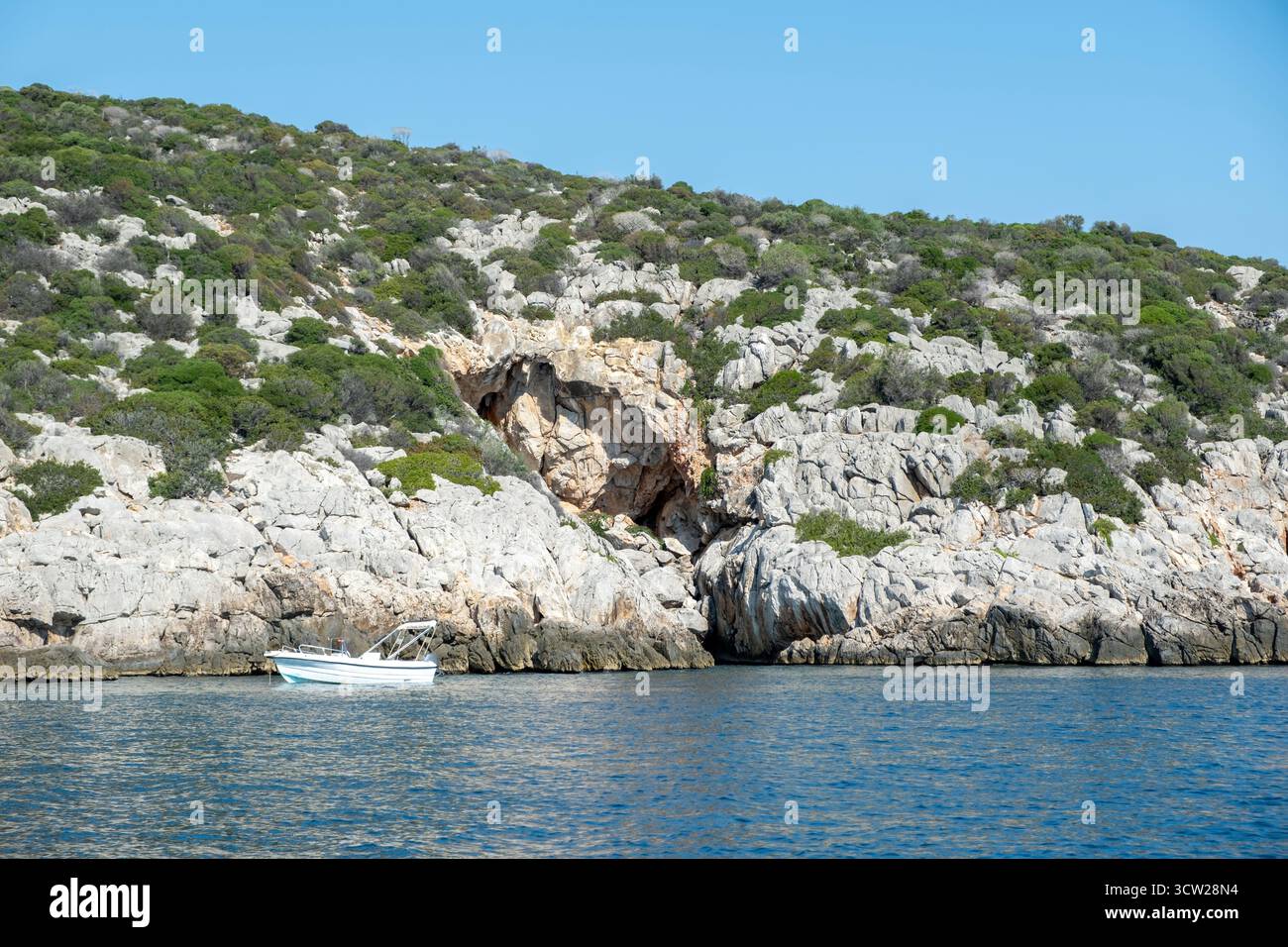 Felsige Küste mit dichtem Grün und einem kleinen Ankerboot in türkisfarbenem Wasser in der Nähe von Tolo, Griechenland. Klarer Tageslicht Himmel Stockfoto