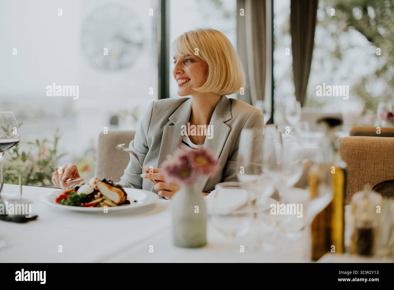 Eine Frau genießt ihr Gourmet-Essen in einem stilvollen Restaurant. Die Szene ist voller edler Esselemente wie Weingläser und frischer Blumen, die einen schaffen Stockfoto