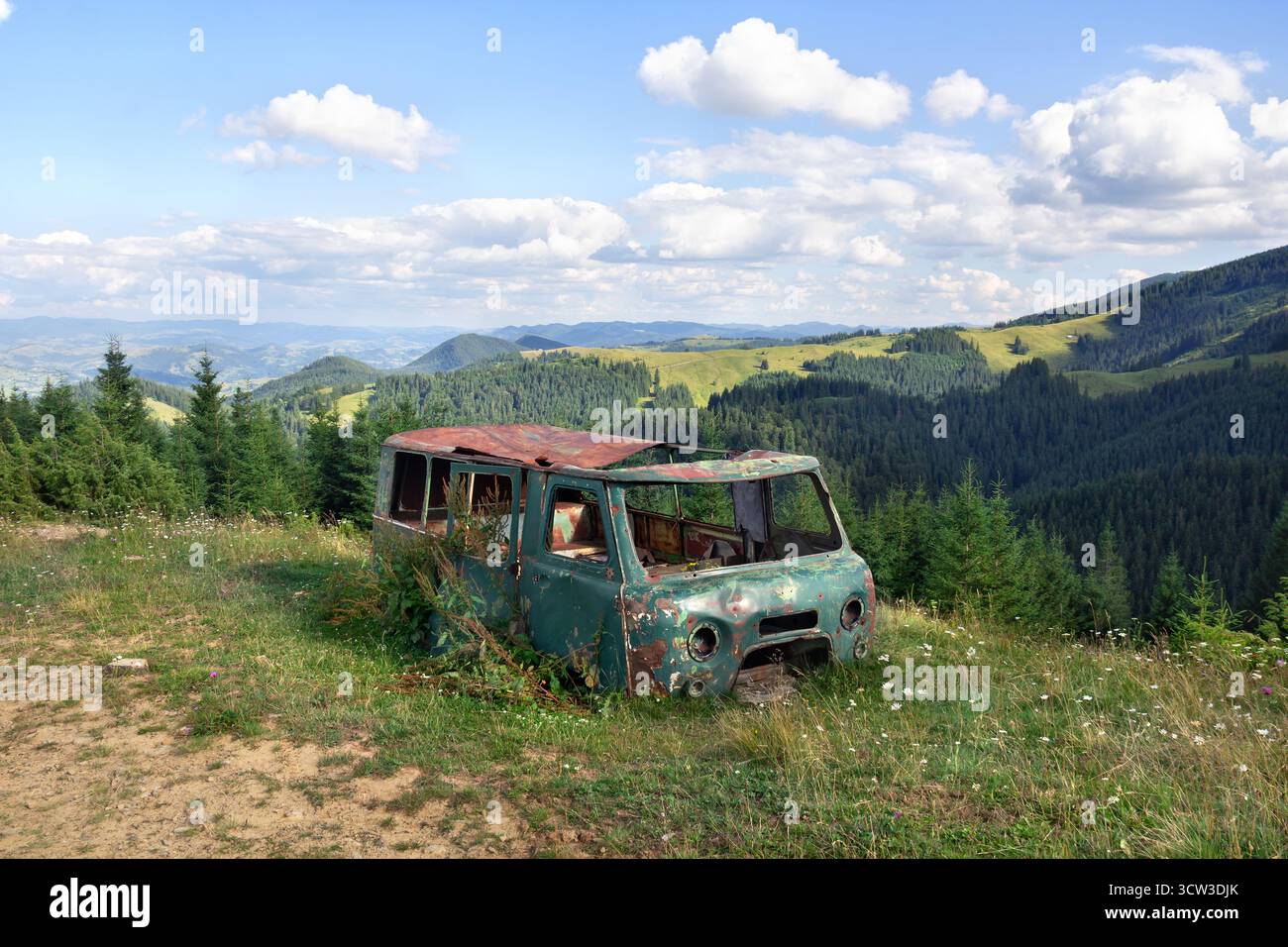 Ein alter rostiger und kaputter Van ohne Fenster oder Scheinwerfer, im Gras zwischen malerischen Bergen verlassen. Oldtimer-Verfall in wilder Natur Stockfoto