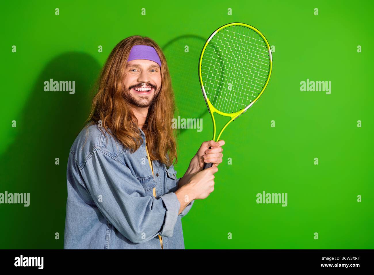 Lächelnder junger Mann, der Tennisschläger auf leuchtendem grünem Hintergrund hält und fröhlichen Retro-Stil zeigt Stockfoto