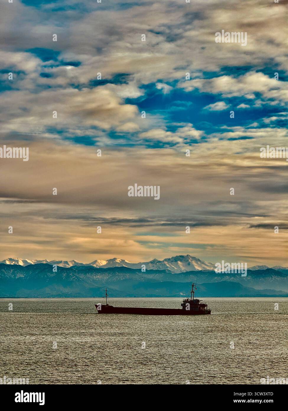 Frachtschiff segelt auf ruhigem Meer mit schneebedeckten Bergen im Hintergrund unter bewölktem Himmel Stockfoto