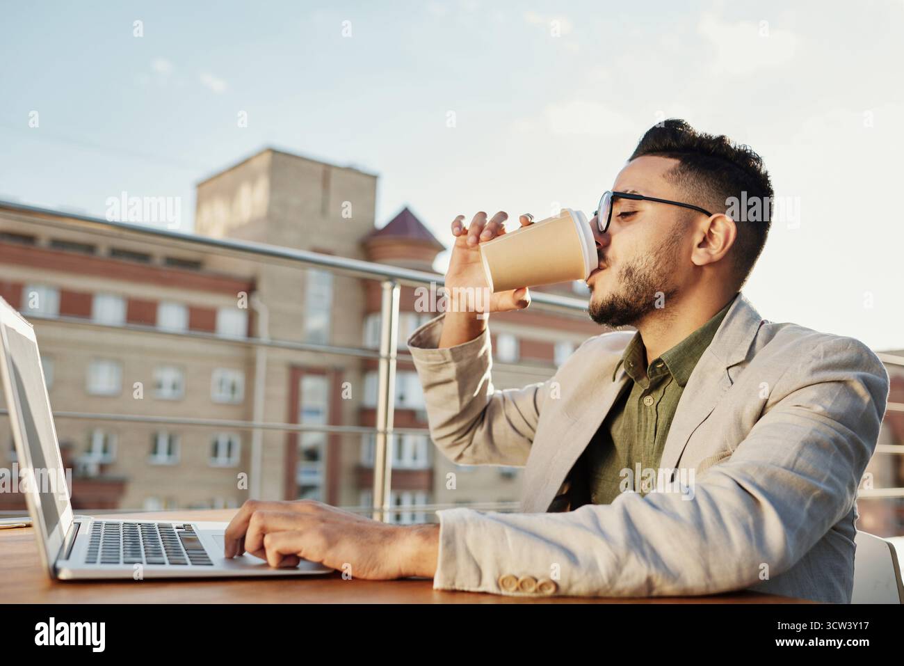 Junger attraktiver Mann aus dem Nahen Osten, der Gläser trägt und Kaffee aus dem Pappbecher weiß trinkt, sitzt am Tisch mit Laptop auf der Dachterrasse des Cafés Stockfoto