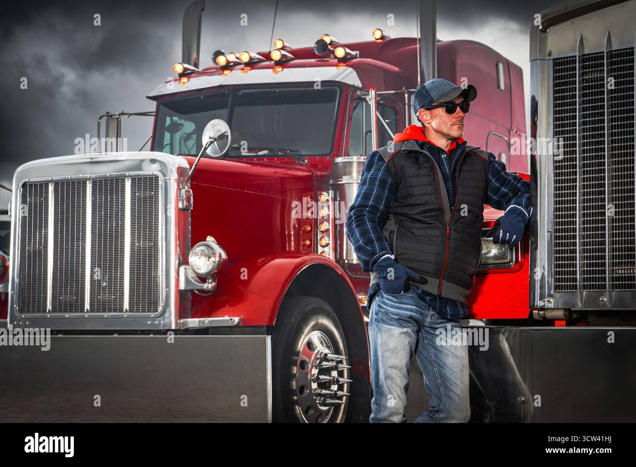 Ein Lkw-Fahrer lehnt sich unter bewölktem Himmel an einen roten Sattelwagen. Die Szene zeigt den Fahrer in lässiger Kleidung und macht tagsüber eine Pause. Stockfoto