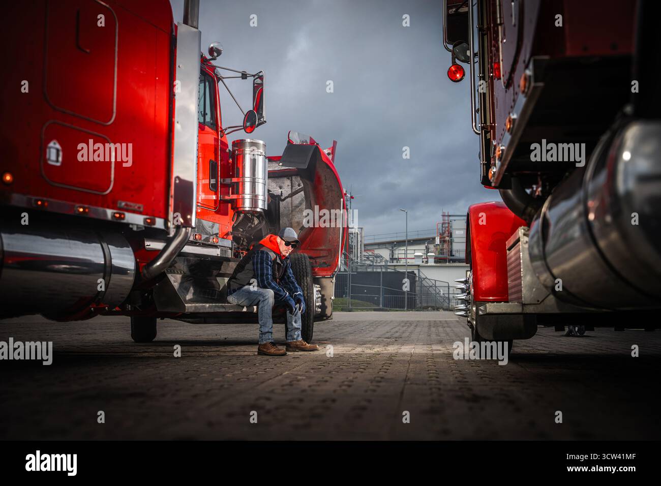 Zwei Lkw-Fahrer sitzen auf den Stufen eines großen roten Lkws und genießen eine Pause in einer industriellen Umgebung, während dunkle Wolken über ihnen hängen. Stockfoto