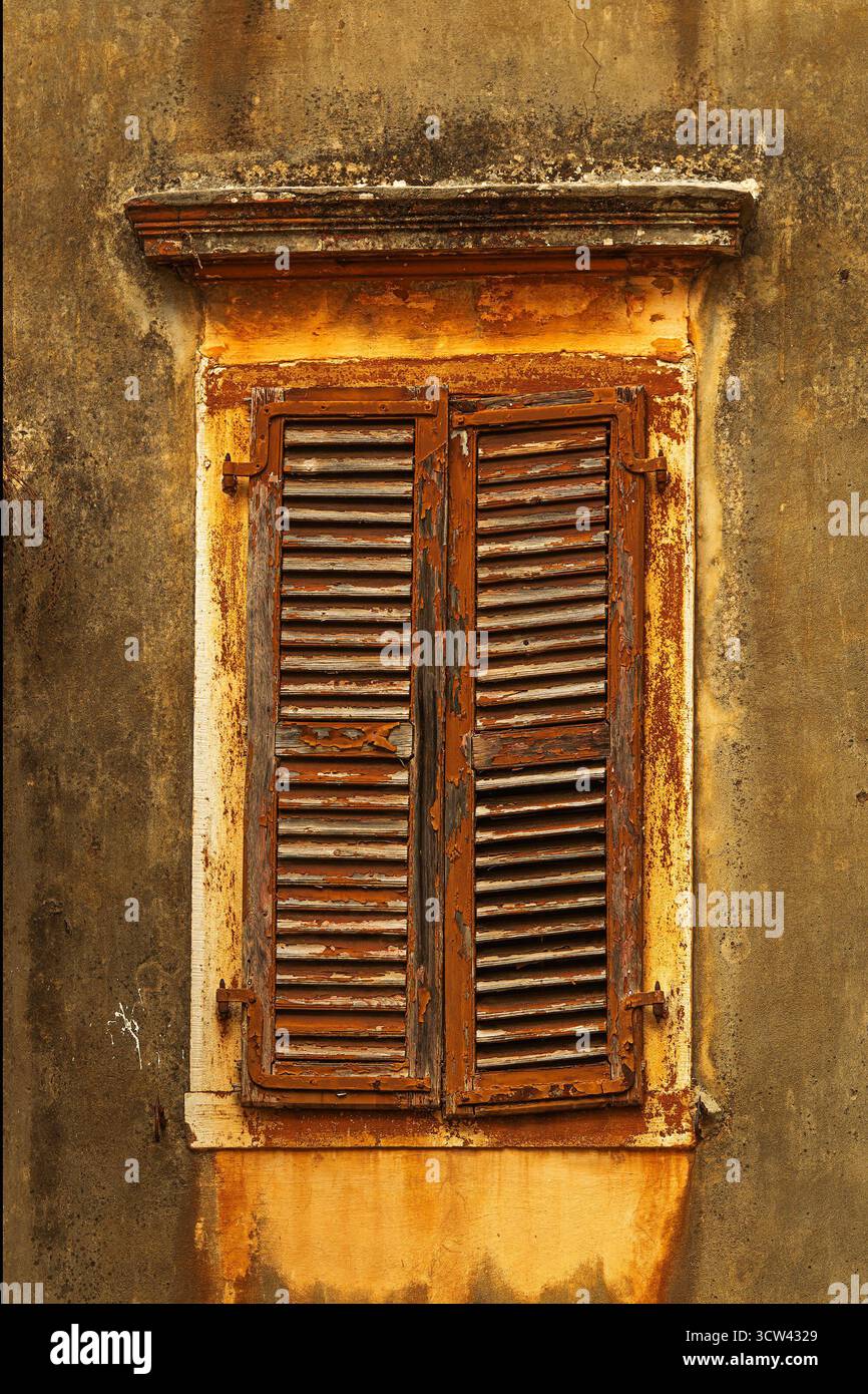 Ein Blick aus der Nähe auf alte Holzläden mit abblätternder Farbe und rostigen Scharnieren an einer strukturierten gelben Wand in Korfu Stadt. Stockfoto