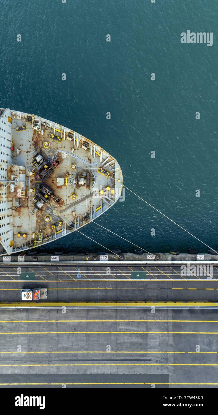 Aus der Vogelperspektive eines Schiffsbogens im Kontrast zum dunklen Wasser, neben einer Brücke mit gelben Linien und einem kleinen Auto, Ports of Auckland, Neuseeland. Stockfoto