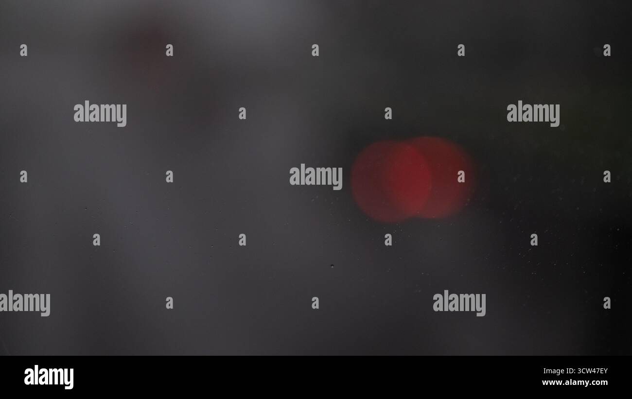 Moody abstrakte Ansicht durch ein nebeliges oder regnerisches Fenster mit verschwommenen roten Lichtern in der Ferne. Atmosphärische urbane Szene mit weichem Fokus und Bokeh-Effekt, Stockfoto