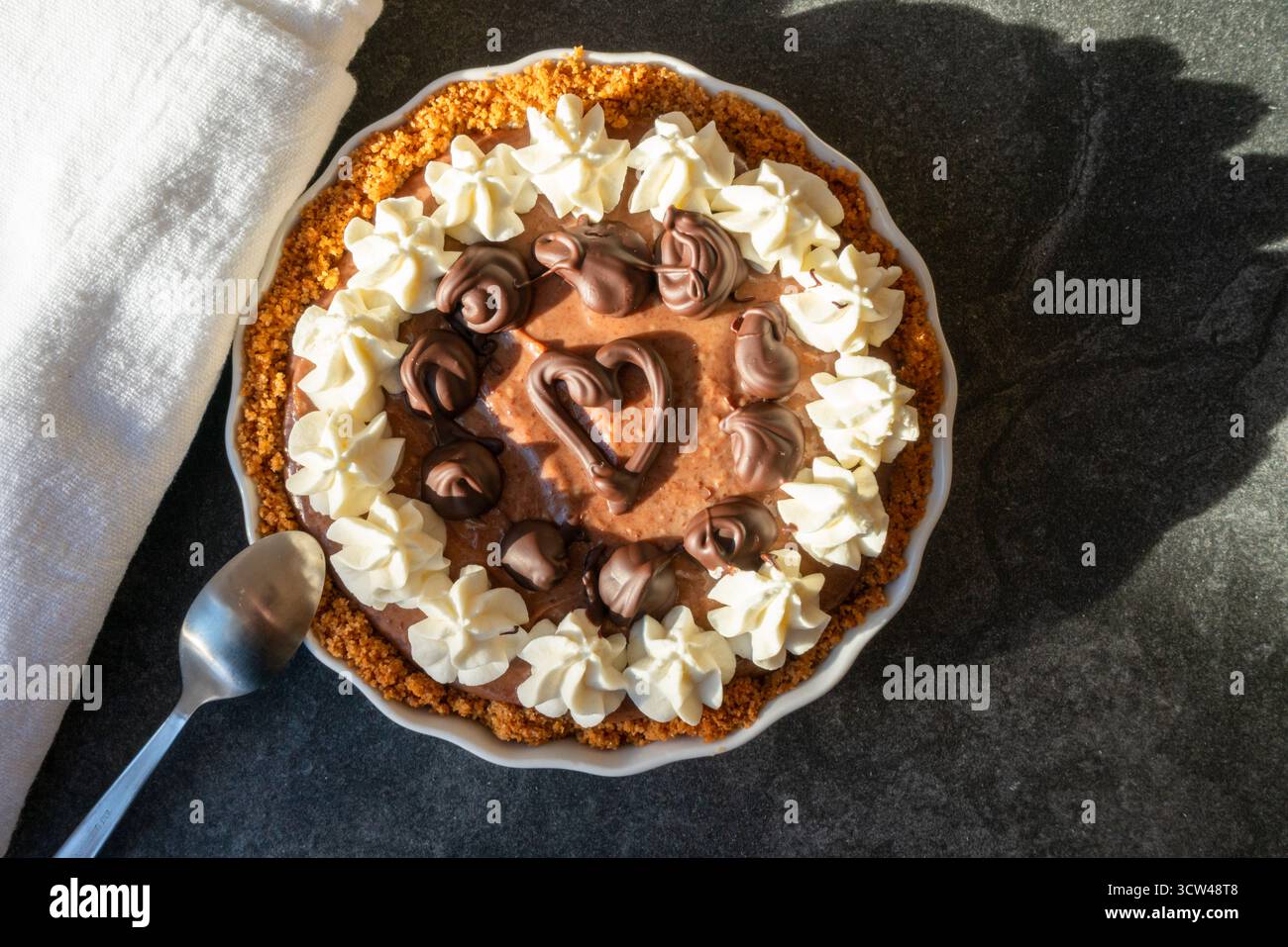 Blick von oben auf einen Schokoladenkuchen mit Schlagsahne und einem Herz in der Mitte, auf einem dunklen Tisch mit einer weißen Serviette in warmem natürlichem Licht. Stockfoto