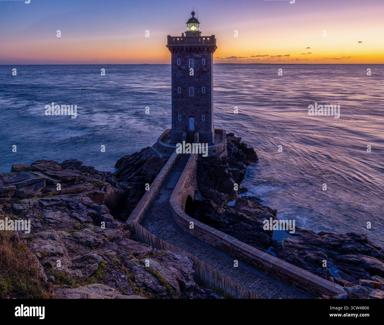 Ein einsamer Leuchtturm, Kermorvan Lighthouse, bewacht die zerklüftete Küste, die in den warmen Farben der untergehenden Sonne getaucht ist. Der Weg zum Leuchtturm Stockfoto