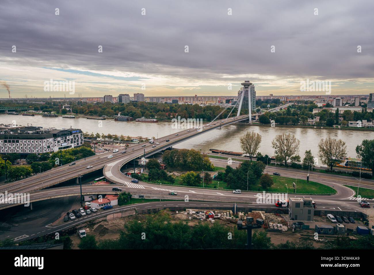 Eine breite Brücke verbindet beide Seiten eines Flusses in der Nähe einer pulsierenden Stadt. Der Verkehr fließt reibungslos, wenn die Herbstbäume die Ufer säumen. Stockfoto