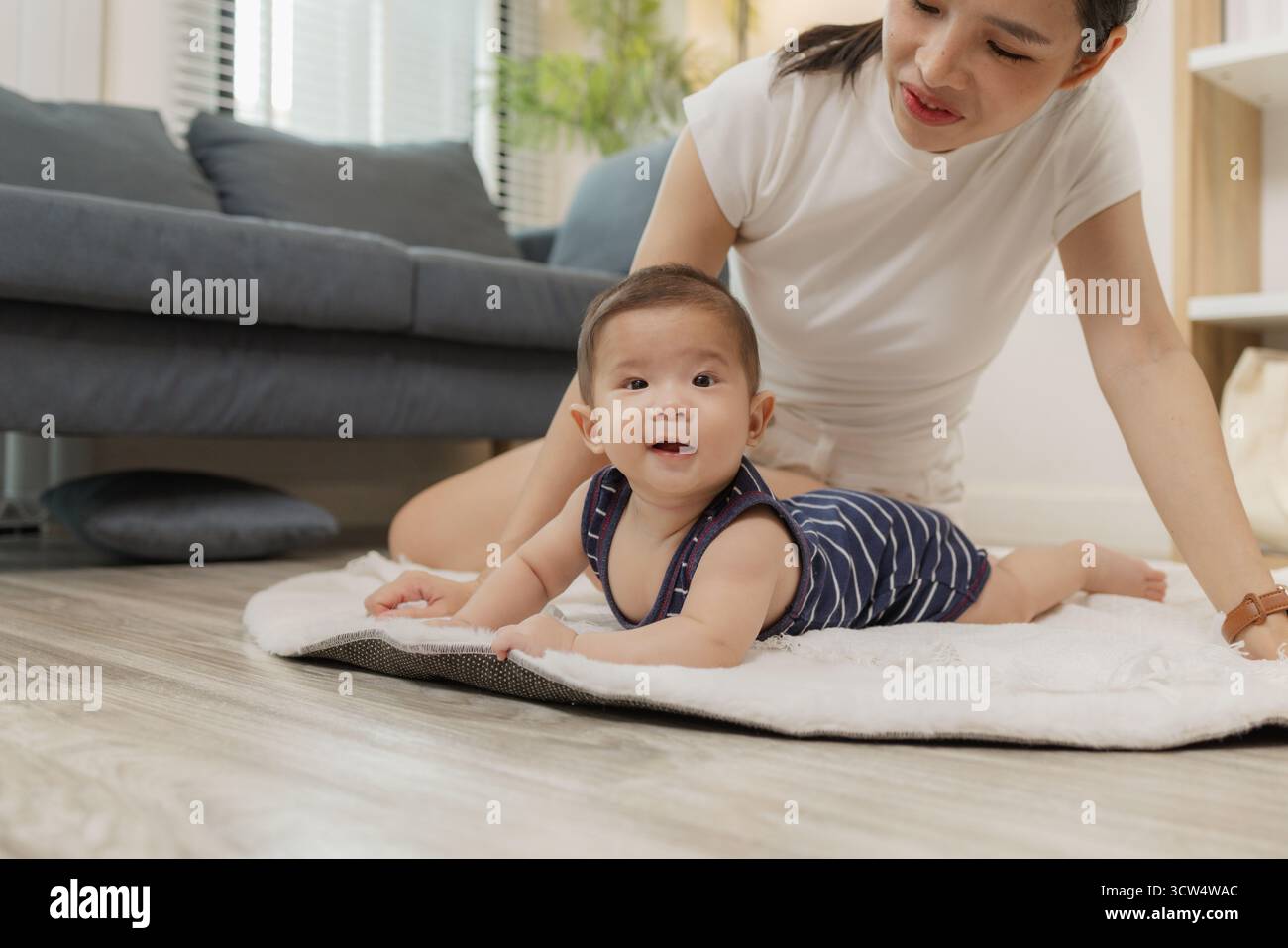 Ein Baby genießt die Bauchzeit auf einer weichen Matte, während die Mutter nah bleibt und es mit einem warmen Lächeln ermutigt. Dieser zärtliche Moment hebt frühe Entwicklungskräfte hervor Stockfoto