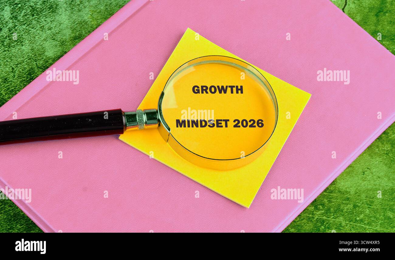 Eine gelbe Haftnotiz mit Growth Mindset 2026 wird auf einem rosafarbenen Notizbuch vergrößert und liegt auf einer grün strukturierten Oberfläche. Stockfoto