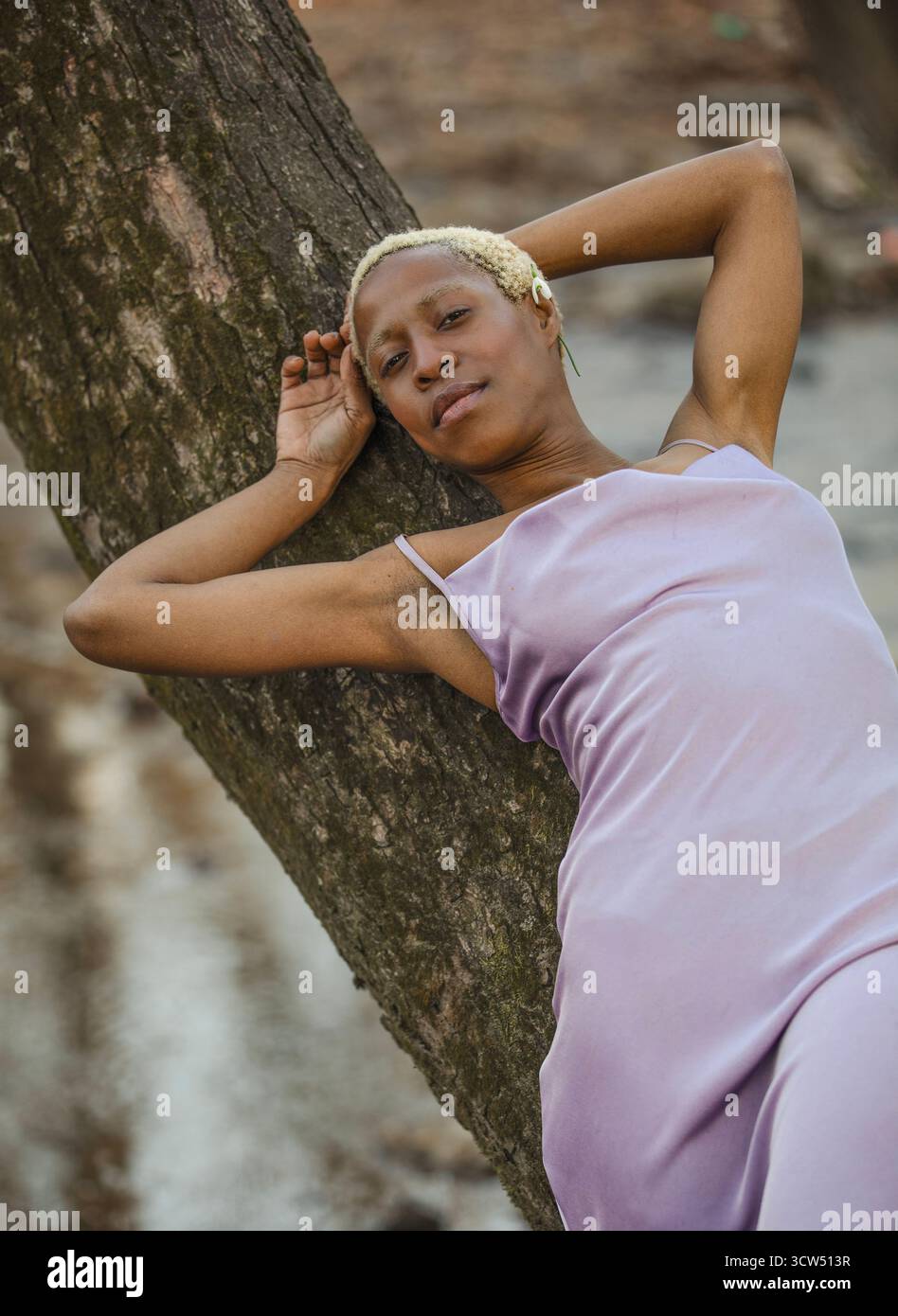 Eine schwarze Frau im Lavendelkleid lehnt sich an einen großen Baum. Im Hintergrund verwischen blattlose Bäume, die den Eindruck einer ruhigen Natur erwecken. Stockfoto