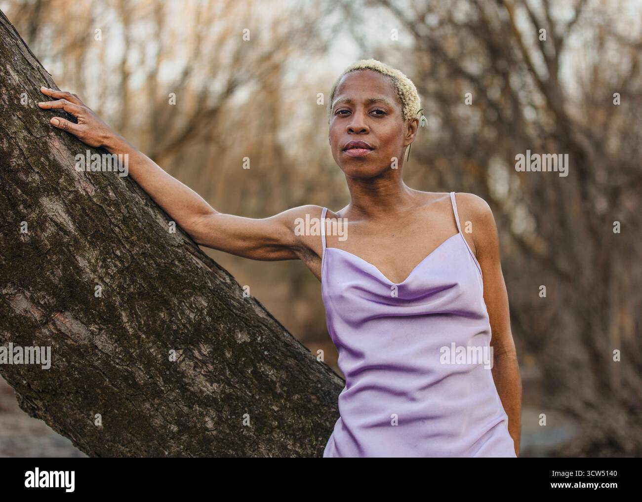 Eine schwarze Frau im Lavendelkleid lehnt sich an einen großen Baum. Im Hintergrund verwischen blattlose Bäume, die den Eindruck einer ruhigen Natur erwecken. Stockfoto