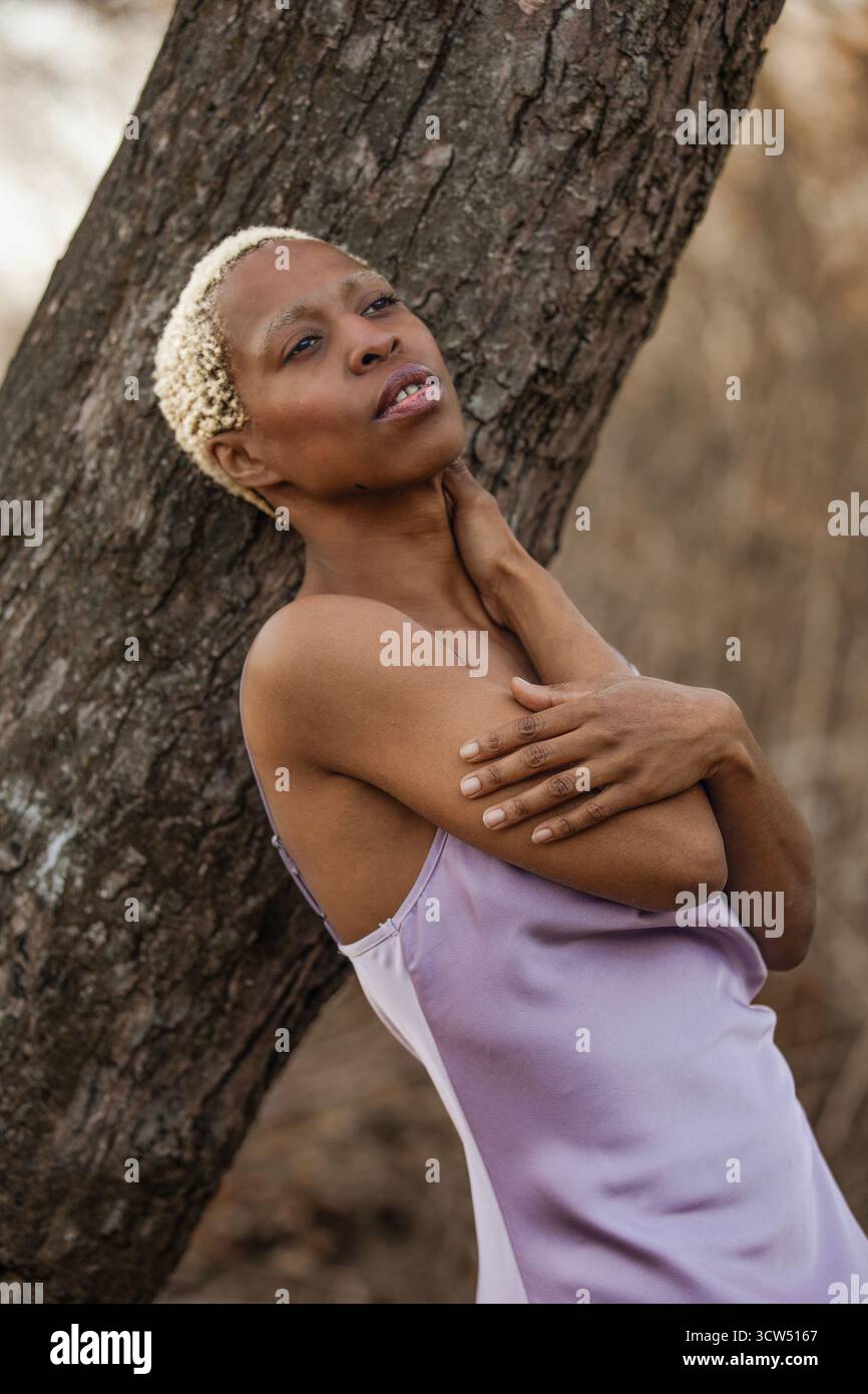 Eine schwarze Frau im Lavendelkleid lehnt sich an einen großen Baum. Im Hintergrund verwischen blattlose Bäume, die den Eindruck einer ruhigen Natur erwecken. Stockfoto