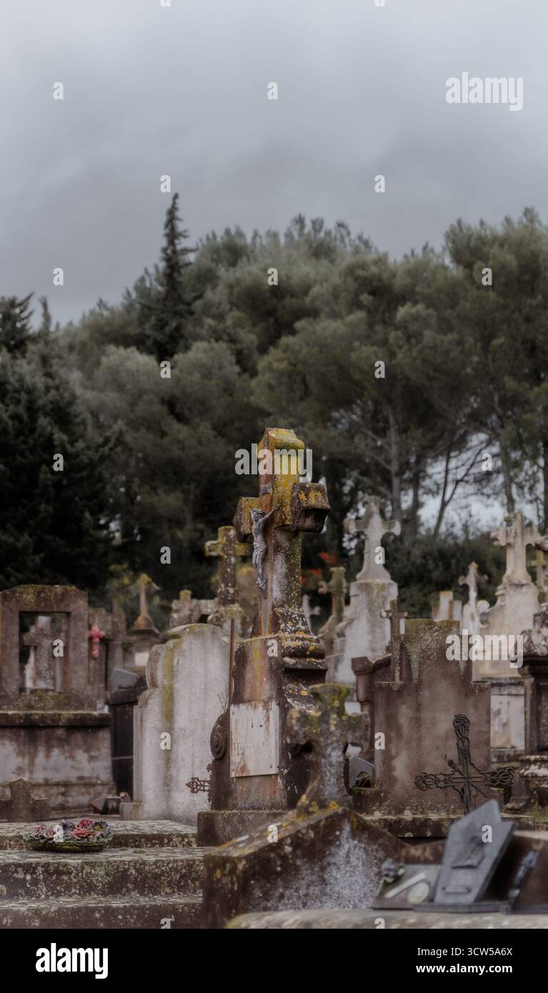 Ein moosbedecktes Kreuz mit einer Figur von Jesus Christus, der auf einem alten Friedhof mit Grabsteinen und Kreuzen an einem grauen, bewölkten Tag gekreuzigt wurde. Stockfoto